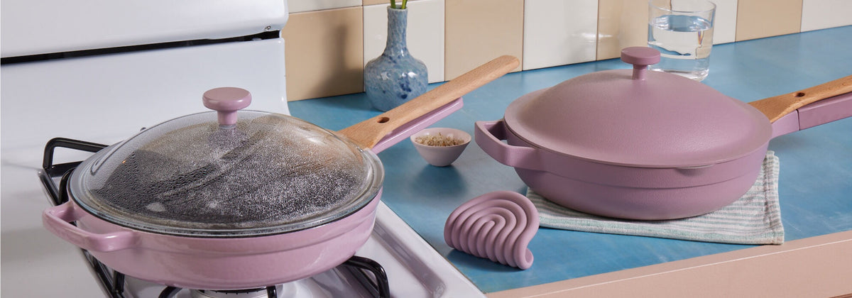 Translation missing: en.Two lavender-colored pans with wooden handles are on a blue kitchen counter; one pan is on a stove with a glass lid, and the other is on a striped cloth beside kitchen accessories and a glass of water