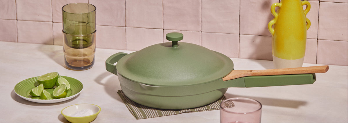 Translation missing: en.A green lidded pan with a wooden handle sits on a placemat. Nearby are a small bowl of salt, a plate of lime wedges, stacked green glasses, a pink glass, and a yellow vase, all on a light countertop with tiled wall