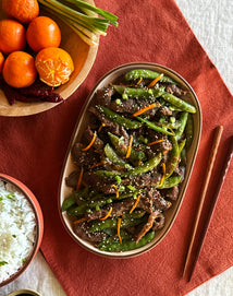 Plated dish of green beans and beef with mandarin oranges and rice on a red placemat.