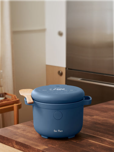A blue electric cooker labeled Our Place sits on a countertop next to a potted plant and a bottle of oil.