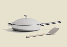 A modern white frying pan with a metal handle and lid sits on a light surface, next to a metal spatula with a light-colored handle.