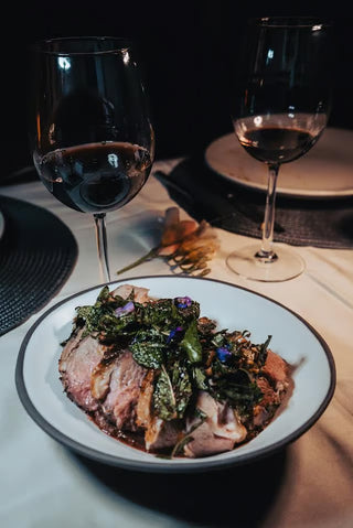 A plate of sliced cooked meat topped with herbs and flowers, set on a table with two glasses of red wine and place settings.