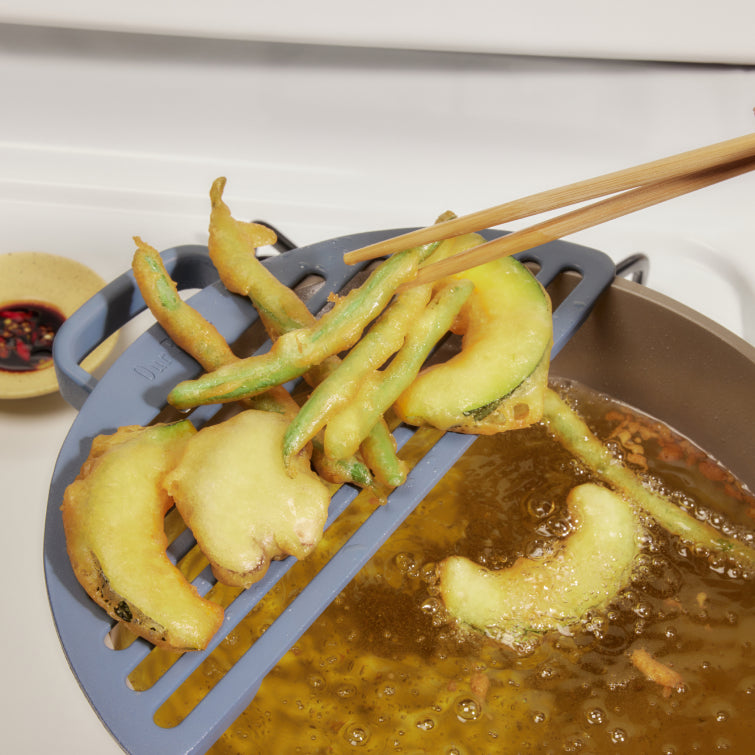 Fried vegetables drip drying on a blue Fry Deck over a pot of oil