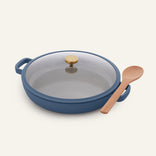 The Dual Handle Always Pan in blue, featuring a nonstick surface, glass lid, and gold knob, is placed next to a wooden spoon on a light background.