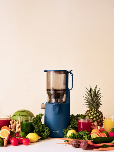 A blue juicer on a kitchen counter with green juice, fresh fruits, vegetables, and a knife. Kitchen utensils and an aloe plant are in the background.