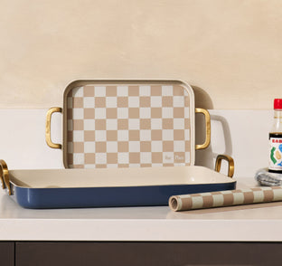Two rectangular baking dishes with gold handles and a beige checkered pattern are on a countertop, alongside a matching roll of paper and a bottle of seasoning.