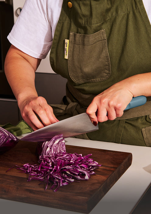 Person wearing a green apron slicing purple cabbage with a large knife on a wooden cutting board.