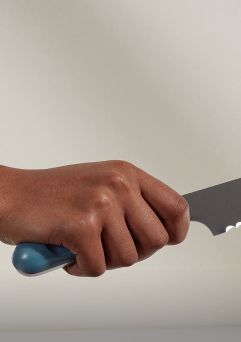 A hand holding a serrated kitchen knife with a blue handle against a plain background.