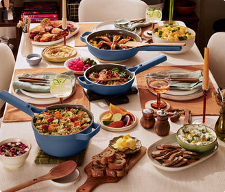 A dining table set with various dishes, including salads, pasta, bread, roasted chicken, dips, and drinks, with blue cookware and neatly arranged place settings.