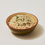 A bowl of cooked brown rice topped with chopped herbs, placed on a light-colored surface.