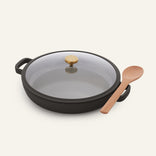 The Dual Handle Always Pan, featuring a glass lid and gold handles in black, is placed next to a wooden spoon on a plain light background.