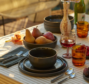 A table set with black dishes, bowls of pears, glasses with drinks, utensils, and a striped tablecloth in sunlight.
