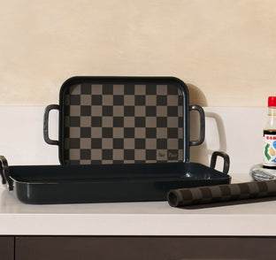 Two black rectangular trays with checkerboard patterns are stacked on a kitchen counter next to a soy sauce bottle and a roll of plastic wrap.