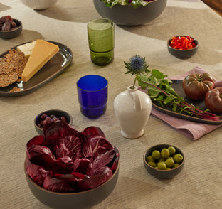A table set with bowls of radicchio, olives, tomatoes, sliced peppers, crackers, cheese, dates, a blue glass, a green glass, and a small white vase with a thistle flower.