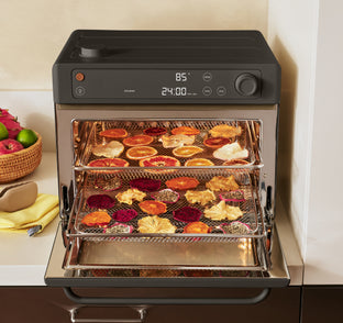 A countertop electric air fryer oven with its door open, showing two racks loaded with assorted dried fruit slices and pieces. The appliance has a black top and control panel with a stainless steel interior and door.