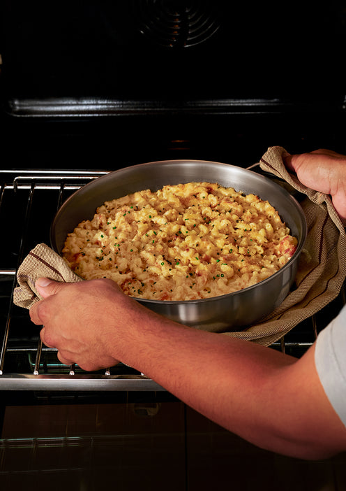A person uses oven mitts to remove a round baking dish of cooked macaroni and cheese from an oven.