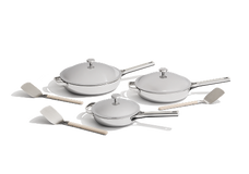 A set of three white lidded pans with metal handles, accompanied by four light-colored cooking utensils, displayed on a white background.