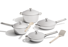 A set of four stainless steel pots and pans with lids, one wooden spoon, and one spatula, arranged on a white background.
