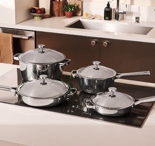 A set of four stainless steel pots and pans with lids is displayed on a kitchen countertop near a sink.