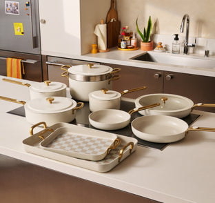A set of white and gold cookware, including pots, pans, and baking dishes, arranged on a kitchen counter and stovetop in a modern kitchen.