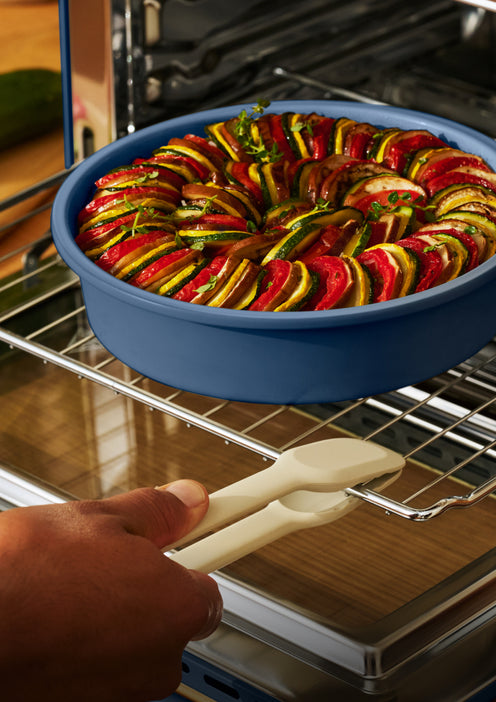 A hand uses kitchen tongs to pull a round blue baking dish filled with sliced ratatouille out of an oven.