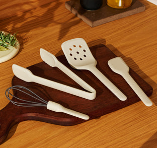 A set of cream-colored kitchen utensils including a whisk, tongs, a slotted turner, and a spatula arranged on a wooden cutting board.