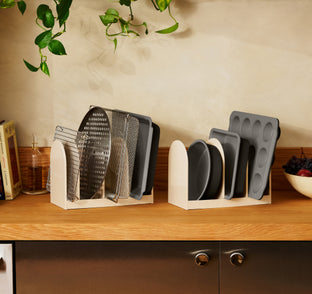 Two light-colored kitchen organizers holding various metal baking sheets, cooling racks, and muffin pans on a wooden countertop.