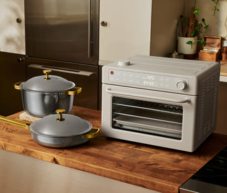 A countertop oven with a digital display sits on a wooden kitchen counter next to two gray pots with gold handles and lids.