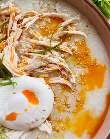 A bowl of congee topped with shredded chicken, a poached egg, sesame seeds, sliced green onions, and drizzled with chili oil.