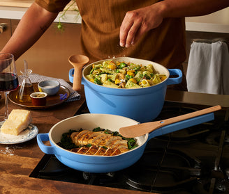 Person cooking pasta and greens in a blue pot on a stove, with a glass of red wine and cheese on the side.