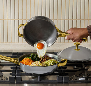 A hand pours a cooked sunny-side-up egg from a small pan into a larger pan containing various shredded vegetables, both pans have gold handles, on a stovetop.