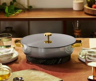 A lidded gray and gold pot sits on a black trivet in the center of a set dining table with plates, glasses, and napkins.