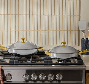 Two covered pans with gold handles are on a stainless steel gas stove; utensils are in a holder on the right, and a tiled backsplash is in the background.