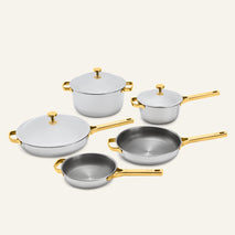 The Titanium Pro Cookware Set+ 2.0 includes a large pot, a saucepan, two frying pans, and a lidded skillet, all featuring silver-toned metal bodies with gold-toned handles and lids.