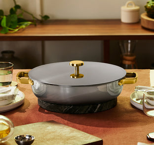 A round, lidded pot with gold handles and a gold knob sits on a marble trivet, surrounded by neatly set dining tableware in a warmly lit room.