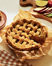 Apple pie with lattice crust on brown paper, surrounded by sliced apples and a wooden cutting board.