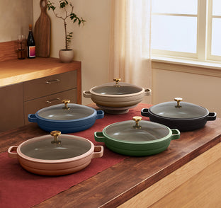 Five shallow, round pans with glass lids and gold handles in different colors are arranged on a kitchen counter with a red runner.