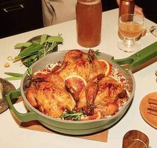 Roasted chicken dish with herbs and oranges in a green skillet on a table with drinks.