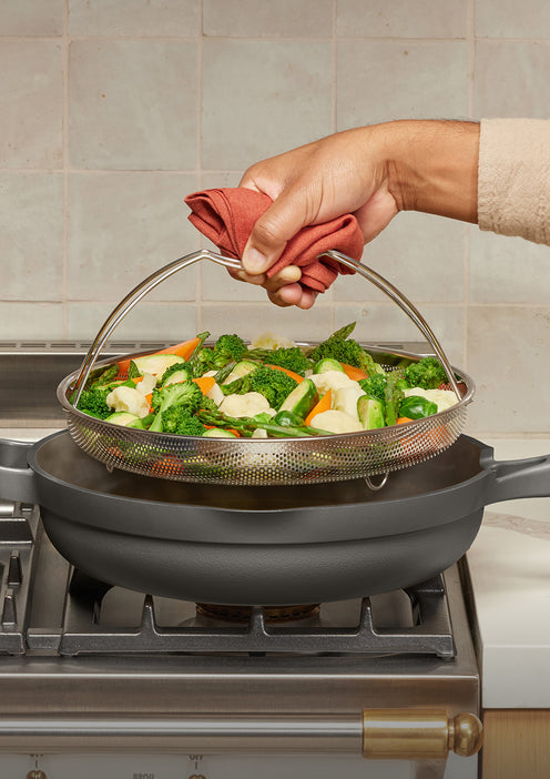 Person using a strainer with vegetables over a frying pan on a stove.