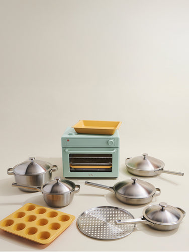 A set of stainless steel cookware, a green mini oven with a yellow tray, a yellow silicone muffin mold, and a round metal grate are arranged on a plain light background.