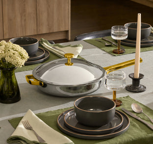 A set dining table with green placemats, gray dishware, a glass vase with flowers, a lit candle, and a covered stainless steel pan with gold handle and knob.