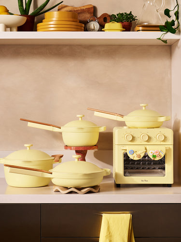 A pastel yellow kitchen set with pots, pans, and a mini oven is displayed on a table surrounded by various foods, including pastries, popcorn, cookies, chicken, burgers, salad, and bread rolls.