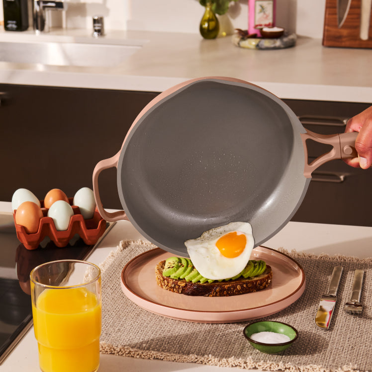 Person lifting a frying pan with an egg on toast, next to a glass of orange juice and avocado toast.