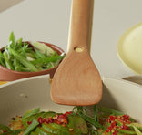 Wooden spatula over a dish of green vegetables with red toppings on a neutral background