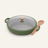 The Dual Handle Always Pan in green, featuring matching handles, a glass lid with a gold knob, and accompanied by a wooden spoon resting beside it, is displayed on a light background.