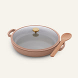 The Dual Handle Always Pan, in a round beige nonstick design with a glass lid topped by a gold knob and two handles, is paired with a wooden spoon and set against an off-white background.