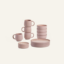 The Dinnerware Set features four pink ceramic bowls, mugs, and plates, all neatly arranged against a plain light background.