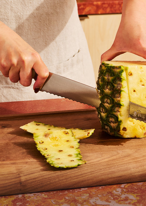 A person slices the skin off a pineapple on a wooden cutting board using a serrated knife.