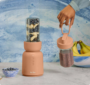A peach-colored blender with a jar of bananas and blueberries inside, a hand holding a jar of blended smoothie, and bananas and a bowl of blueberries on the counter.