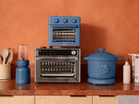 Kitchen appliances including a blue toaster oven, blender, and pot on a wooden counter with an orange wall background.